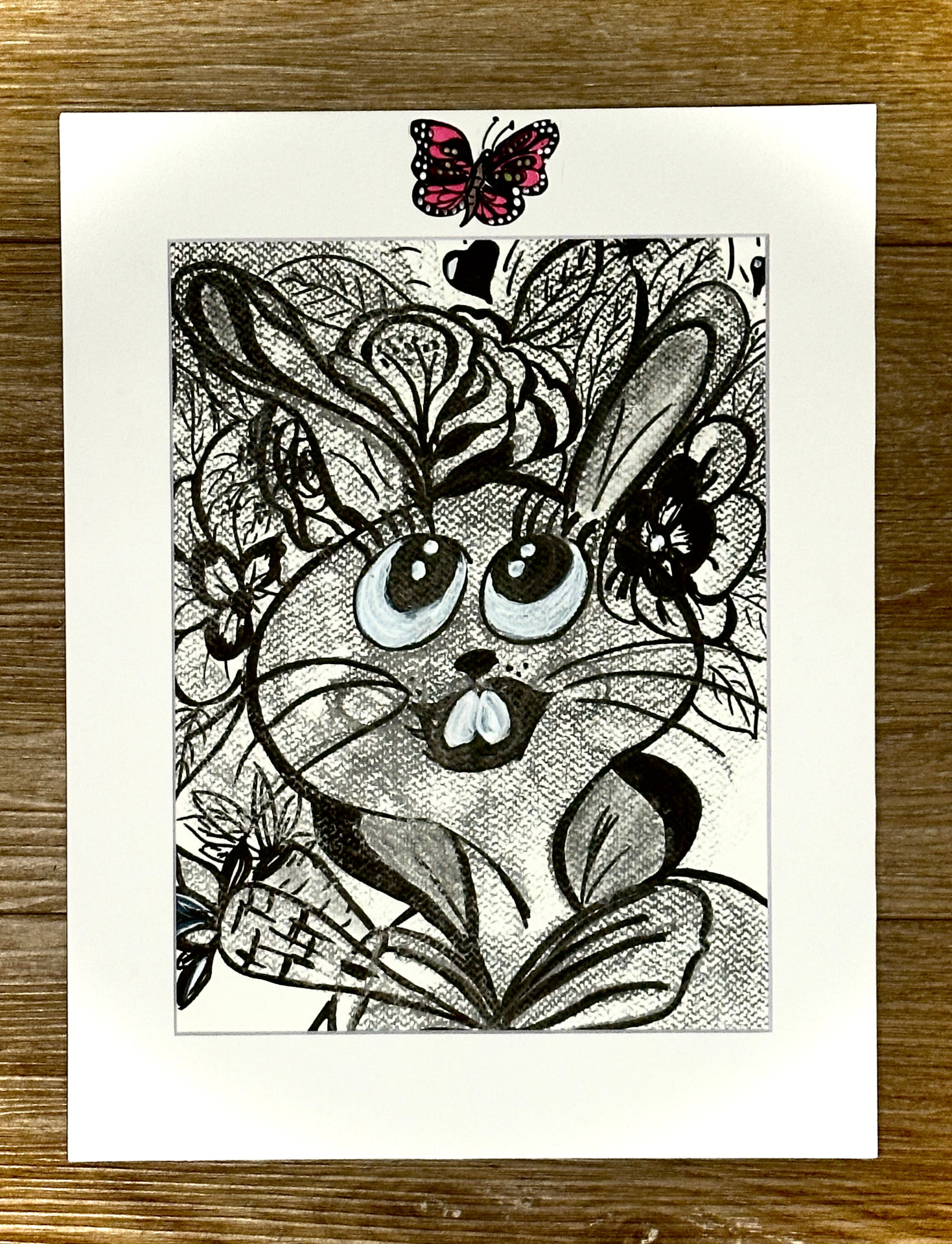 A hand drawn ink and charcoal illustration of a big-eyed bunny surrounded by flowers and leaves on textured paper, matted in white with a small hand-painted pink butterfly on the mat, by Céline